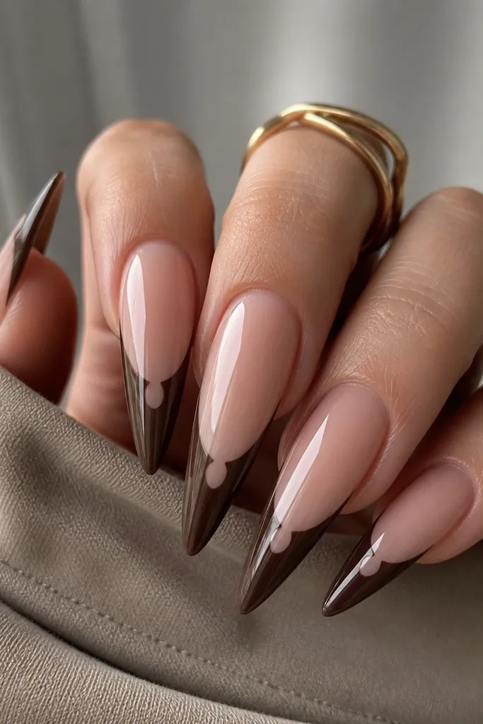 Long stiletto nails with a sheer nude base. The French tips are chocolate brown, but there's a tiny nude window in the center of the smile line, creating a negative-space notch. The brown looks matte-to-satin with a glossy top coat on top.